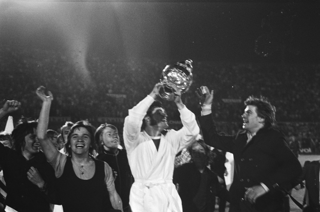 Eine Gruppe von Menschen feiert mit einem Pokal, die zentrale Figur hält ihn triumphierend hoch, während andere jubeln und klatschen, vor einem hell erleuchteten Hintergrund in Schwarz-Weiß.