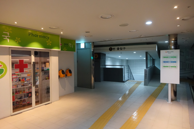 Eingang zu einer Apotheke mit einem "Apotheke"-Schild auf der linken Seite, einer Tafel mit Text auf der rechten Seite und beleuchteten Lampen über dem Dach.