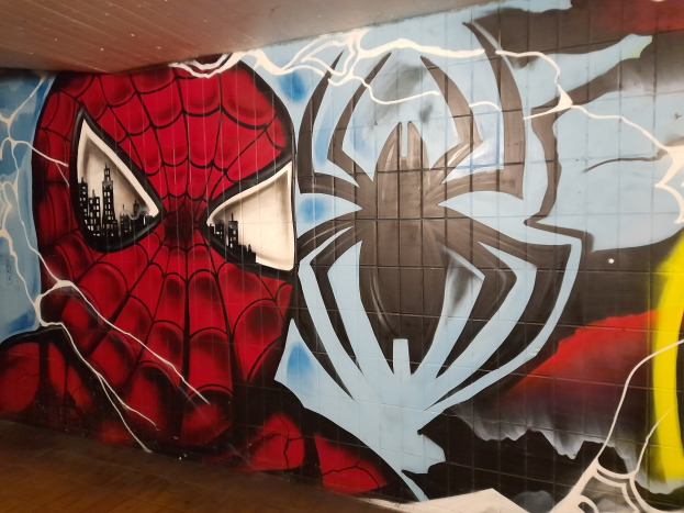 Buntes Wandgemälde von Spider-Man in rot und blauem Kostüm mit Webmuster und großem Spider-Logo, positioniert an einer Gebäudewand zwischen Boden und Decke, mit großen Augen und leicht geöffnetem Mund.