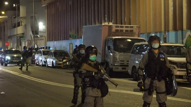 Eine Gruppe von Polizisten in Helmen, Handschuhen und Gesichtsmasken steht nachts auf einer Straße, einige halten Waffen, mit Fahrzeugen, Laternen, Schildern und Gebäuden im Hintergrund.