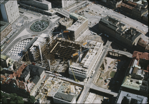 Luftaufnahme einer Stadt mit einem großen Gebäude in der Mitte im Bau, umgeben von anderen Gebäuden, Straßen mit Fahrzeugen und Bäumen; Text unten.