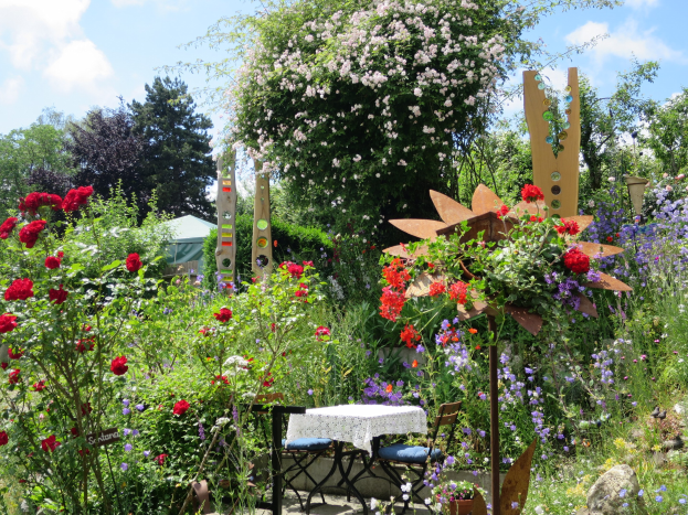 Ein farbenfroher Garten auf der Chelsea Flower Show mit bunten Blumen und Pflanzen, einem Tisch und Stühlen in der Mitte, die mit einem Tuch bedeckt sind, umgeben von Bäumen unter einem bewölkten Himmel.