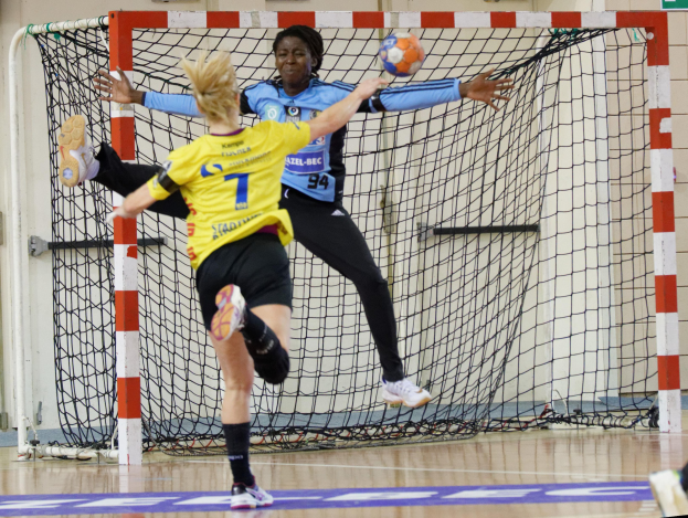 Zwei Frauen beim Handballspielen mit einem Ball in der Luft vor einem Tor, ein Bein rechts sichtbar, beide tragen Schuhe.
