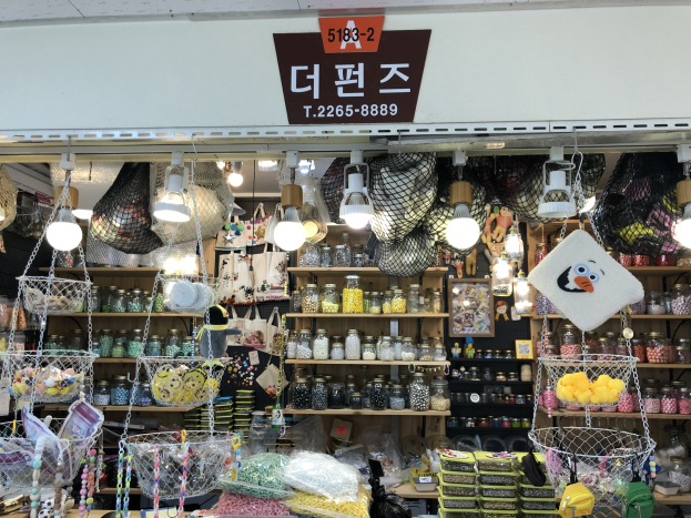 Ein unordentlicher Laden in Seoul mit verschiedenen Gegenständen wie Körpchen, Lichtern, Flaschen und Spielzeugen auf Regalen, mit einer Tafel öber, die anzeigt, dass es einer der besten koreanischen Läden ist.