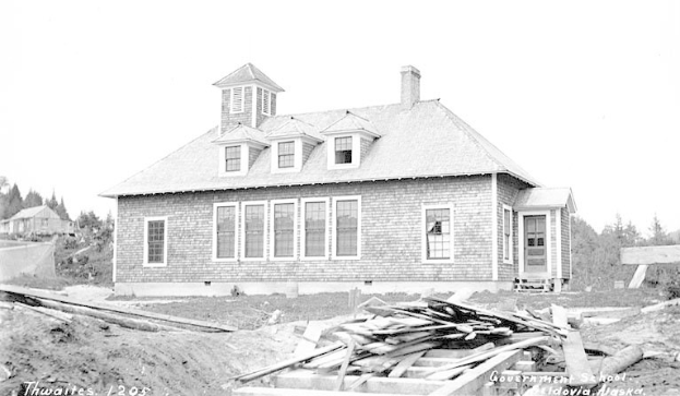 Schwarz-weiß-Foto einer Regierungsgrundschule von 1905, umgeben von Bäumen unter einem klaren Himmel, mit Holzplanken und dem Text "Regierungsgrundschule, Thwaites, 1905" unten.