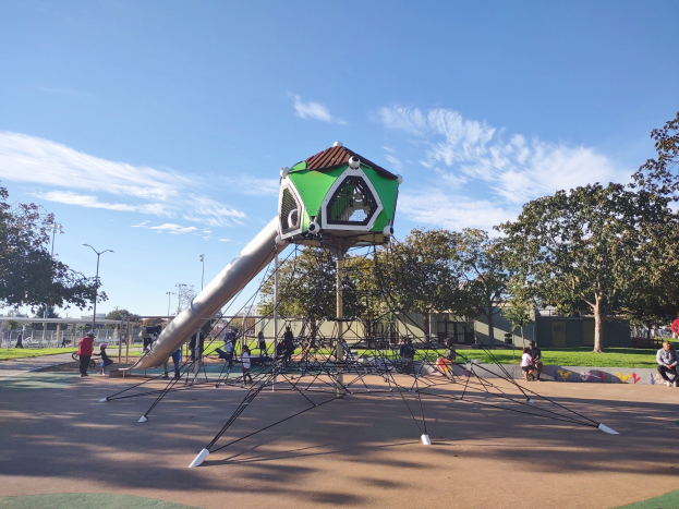 Spielplatz mit einer grün-weißen Spielstruktur, Menschen beim Spielen auf dem Boden, Bäumen, Laternenmasten, Gebäuden und einem bewölkten Himmel im Hintergrund.