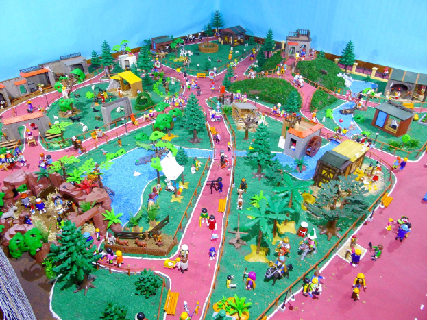 Ein Miniaturmodell eines Zoos mit zahlreichen Besuchern, umgeben von Bäumen, Häusern, Pflanzen, Gras, einem Zaun und anderen Elementen, vor einer Hintergrundwand mit Text unten.