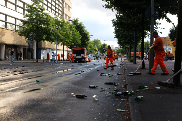 Eine Gruppe von Menschen in orangenen Uniformen sammelt Müll von einer Straße, die mit Flaschen und Schutt übersät ist, mit Bäumen, Pfählen und Fahrzeugen im Hintergrund unter einem bewölkten Himmel.