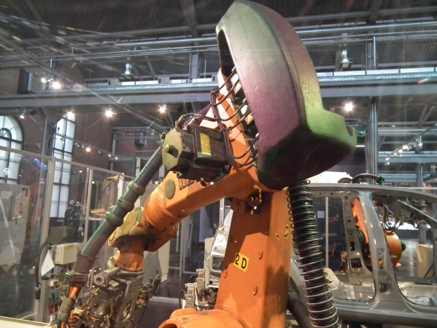 Roboterarm beim Zusammenbau eines Autos in einer industriellen Fabrikumgebung mit Metallstangen, Beleuchtung und anderen Maschinen, Glaswand links, zusätzliche Objekte im Hintergrund.