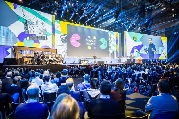 Eine große Menschenmenge, die auf Stühlen vor einer Bühne sitzt, mit einigen Personen, die auf dem Podium in der Vordergrund stehen, einem Tisch mit verschiedenen Gegenständen links und Bildschirmen und Lichtern im Hintergrund auf der Deutschen Internet-Konferenz 2019.