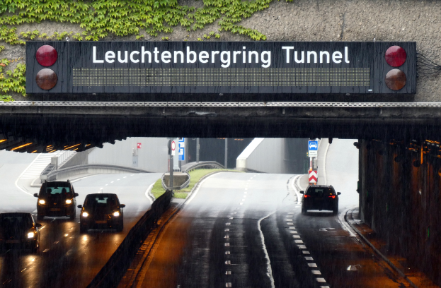 Ein Tunnel mit Autos, die an einem regnerischen Tag hindurchfahren, mit einer Brücke mit einer Texttafel oben drauf, Fahrzeugen auf der Straße unten, einer Wand auf der rechten Seite und Schildern, Pfosten, Gras und Pflanzen im Hintergrund.