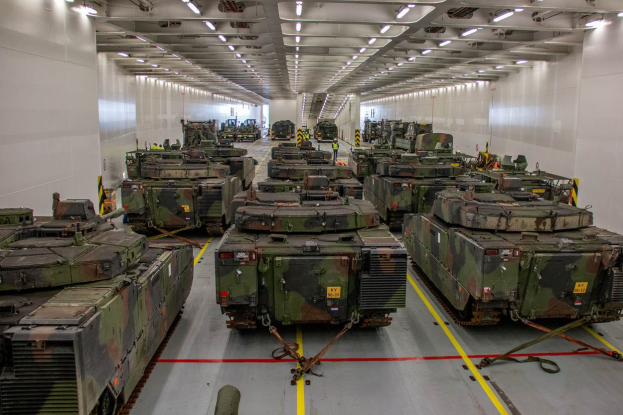 Ein grosser Militaraerraum mit zahlreichen in Reih und Glied aufgestellten Panzer, beleuchtet von Deckenleuchten und von Waenden umgeben.