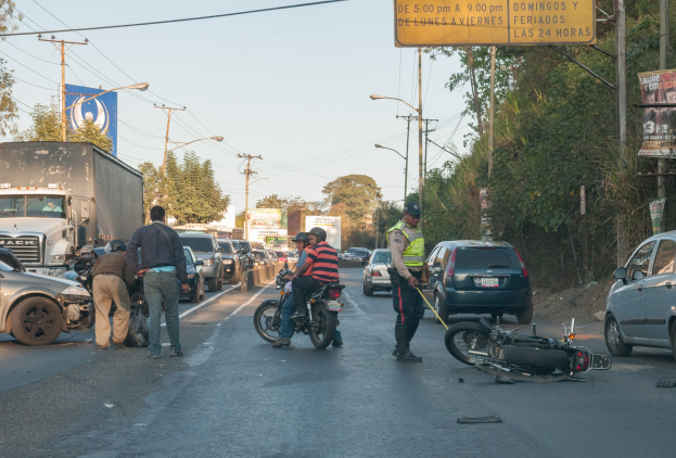 Eine Gruppe von Menschen steht in der Nähe eines verunglückten Motorrads an der Straße mit mehreren Fahrzeugen, Bäumen, Polen und Lichtern im Hintergrund.