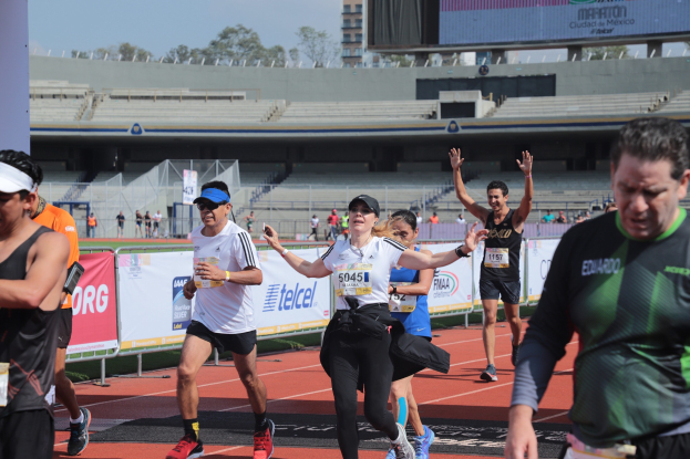 Gruppe von Läufern auf einer Außenbahn in einem Stadion mit Bannern, einer Anzeigetafel, einem Gebäude, Bäumen und einem bewölktem Himmel; einige Läufer tragen Turnschuhe und Hüte.