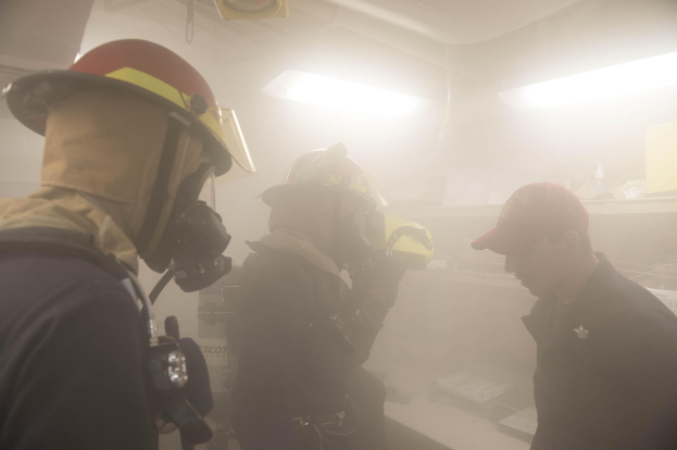 Feuerwehrleute mit Helmen und Rauch aus dem Mund, einer hält eine Kamera, während einer Übung mit Lichtern und Gegenständen im Hintergrund.