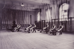 Schwarzes und weißes Bild einer Gruppe von Menschen, die Pilates-Übungen in einem Fitnessstudio mit einer Wand im Hintergrund, Fenstern auf der rechten Seite und Deckenlampen durchführen.