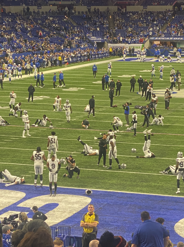 Ein Fußballspiel wird in einem Stadion gespielt, während Menschen auf dem Rasen stehen und einige auf Stühlen im Hintergrund sitzen, während eine Person eine Kamera hält, was auf eine Live-Übertragung hinweist.