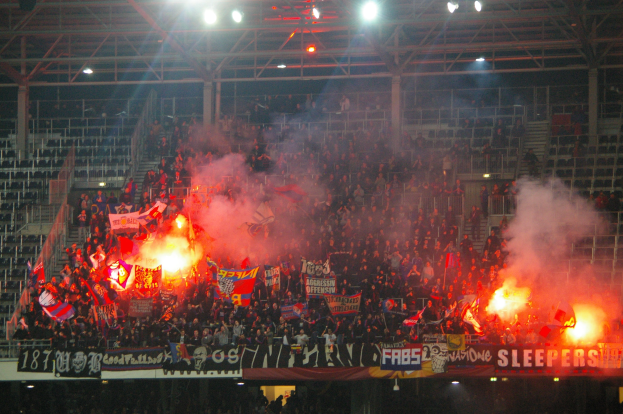 Eine große Menge Menschen in einem Stadion hält Fahnen und Banner, mit Leuchtraketen und Rauch, der von Metallrahmen und Deckenleuchten über ihnen aufsteigt.