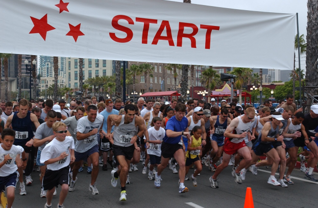 Gruppe von Läufern bei einem Marathon, die an einer Verkehrskegel vorbeiläuft, mit einem Banner und städtischer Umgebung unter einem klaren Himmel.
