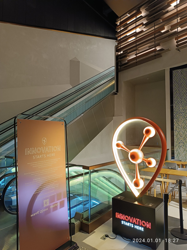 Großes Schild in einem Gebäude mit der Aufschrift "Innovation beginnt hier" und einer Rolltreppe im Hintergrund, Tische und Stühle rechts und eine Tafel links, die auf die HSTP20 Expo hinweist.