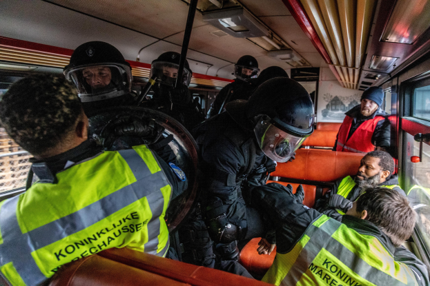 Polizisten in Einsatzausrüstung auf einem Bus sitzend, mit einer Person in der Mitte, sichtbaren Fenstern auf der rechten Seite und einem Plakat an der Wand im Hintergrund.
