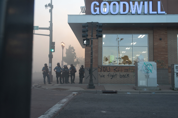 Polizeibeamte in Helmen und taktischer Ausrüstung stehen vor einem gutartigen Geschäft mit Glaswänden, halten Waffen und haben Verkehrssignale und Laternen im Vordergrund und Bäume unter einem klaren Himmel.