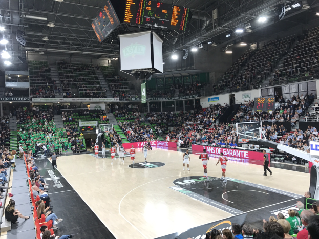 Basketballspiel zwischen Asvel und Galatasaray in einer großen Arena mit Zuschauern, Anzeigetafeln mit den Teamnamen und Überdachung mit Leuchten und Bildschirmen.