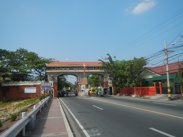Eine Straße mit fahrenden Autos, ein Fußweg mit Geländern auf der linken Seite, Bäume und Gebäude auf beiden Seiten der Straße, ein Tor mit einem Namensschild, Strommasten mit Überlandleitungen und ein bewölkter Himmel im Hintergrund.