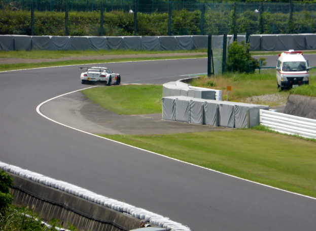 Ein Rennauto, das auf einer Rennstrecke fährt, mit einem Polizeiauto im Hintergrund, umgeben von Gras, Pflanzen, Bäumen und Absperrungen, mit einem Fahrzeug auf der rechten Seite und Text unten.