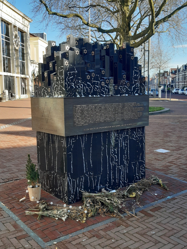 Denkmal in einem Stadtplatz für Holocaust-Opfer mit einem Baum davor, einem Topfgewächse auf dem Boden, verstreuten trockenen Blättern und Gebäuden, Fahrzeugen, Masten und Bäumen im Hintergrund.