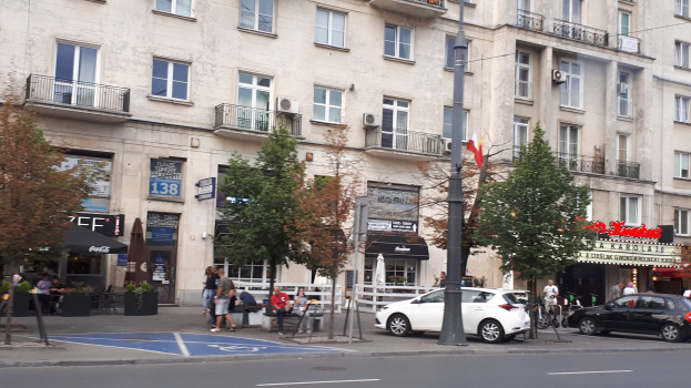 Stadtstraße mit parkenden Autos, Bäumen, einem Gebäude mit Fenstern, Fußgängern, Bänken, Informationsschildern und einem Fahnenmast.