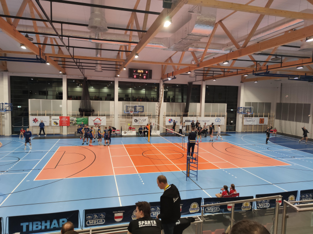 Gruppe von Menschen, die Handball in einer Turnhalle mit Zuschauern spielen, eine Leiter in der Mitte, Geländer, Tafeln mit Text unten und Wände, Säulen, einen Bildschirm und Deckenleuchten im Hintergrund.