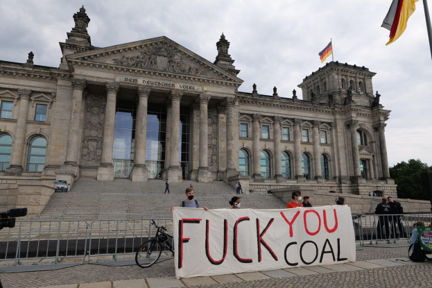 Protestierende halten ein "Fuck You Coal"-Schild vor dem Reichstag in Berlin, mit Bäumen, einer Flagge und einem bewölkten Himmel im Hintergrund.