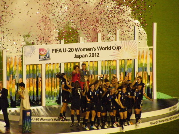 Frauenfußballspielerinnen in schwarzen Trikots feiern auf einer Bühne mit Publikum und einem Hintergrundplakat.