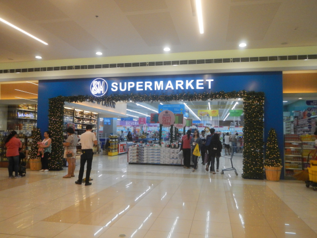 Eine Gruppe von Menschen vor einem Supermarkt, einige mit Einkaufstaschen, Einkaufswagen, Topfpflanze, Regalen mit Gegenständen, Schildern und einer Decke mit Deckenleuchten.