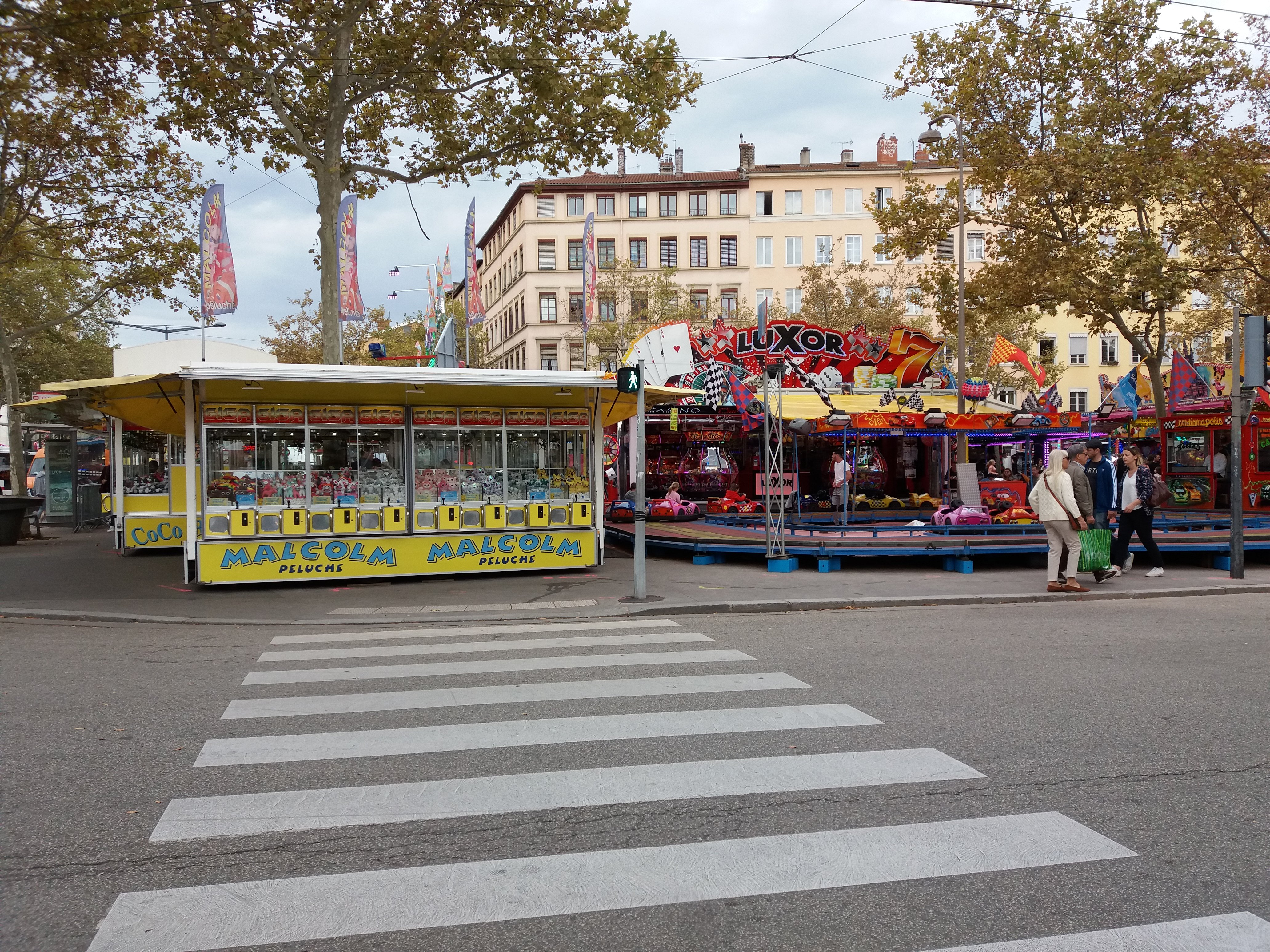 Eine belebte Stadtstraße mit Zebrastreifen, Karussell, Fußgängern, Ständen, Bäumen, Gebäuden, Bannern, Laternenmasten und einer Ampel unter einem bewölkten Himmel.
