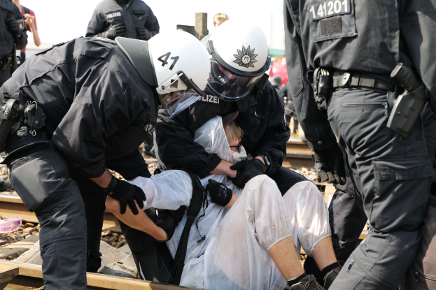 Eine Gruppe von Polizisten in schwarzer Einsatzkleidung verhaftet einen Mann in einem weißen Kleid auf den Gleisen, während andere Menschen in der Nähe stehen und der Himmel im Hintergrund zu sehen ist.