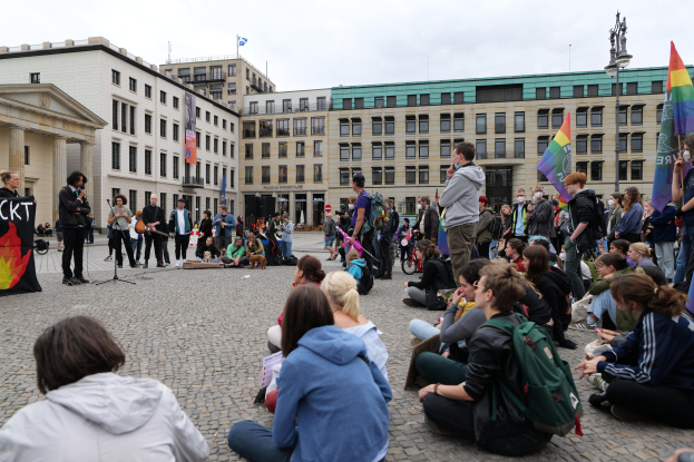 Eine Gruppe von Menschen, die auf dem Boden vor einer Menge mit Fahnen und Spruchbändern sitzt, mit einer Person, die in ein Mikrofon spricht, einer Statue und Gebäuden im Hintergrund bei einer Homo-Hass-Demo in Berlin.