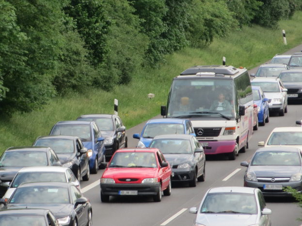Ein Stau auf einer Autobahn mit mehreren Autos und einem Van, Menschen in den Fahrzeugen und Bäume und Gras im Hintergrund.