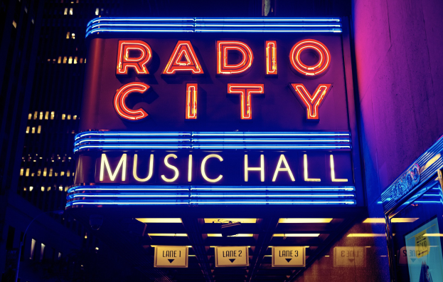 Hell erleuchtetes Radio City Music Hall in New York City mit hohen umliegenden Gebäuden und einem "Radio City"-Schild auf der rechten Seite mit zusätzlichen Schildern darunter.