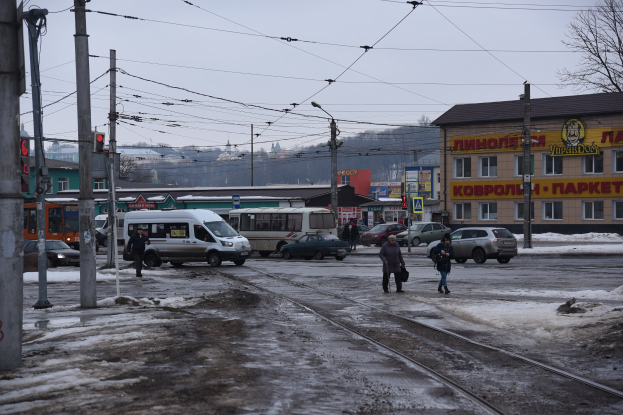 Gruppe von Menschen, die eine Straße neben einer Bahnschiene mit Fahrzeugen, Strommästen, Ampeln, Gebäuden, Bäumen und einem Lieferwagen im Hintergrund überqueren.