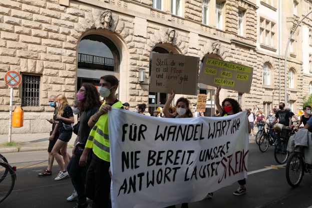 Eine Gruppe von Menschen marschiert bei einer Demonstration auf einer Stadtstraße, einige halten Schilder und andere fahren Fahrräder, mit einem historischen Gebäude mit gewölbigen Fenstern, Pfeilern und Skulpturen im Hintergrund.