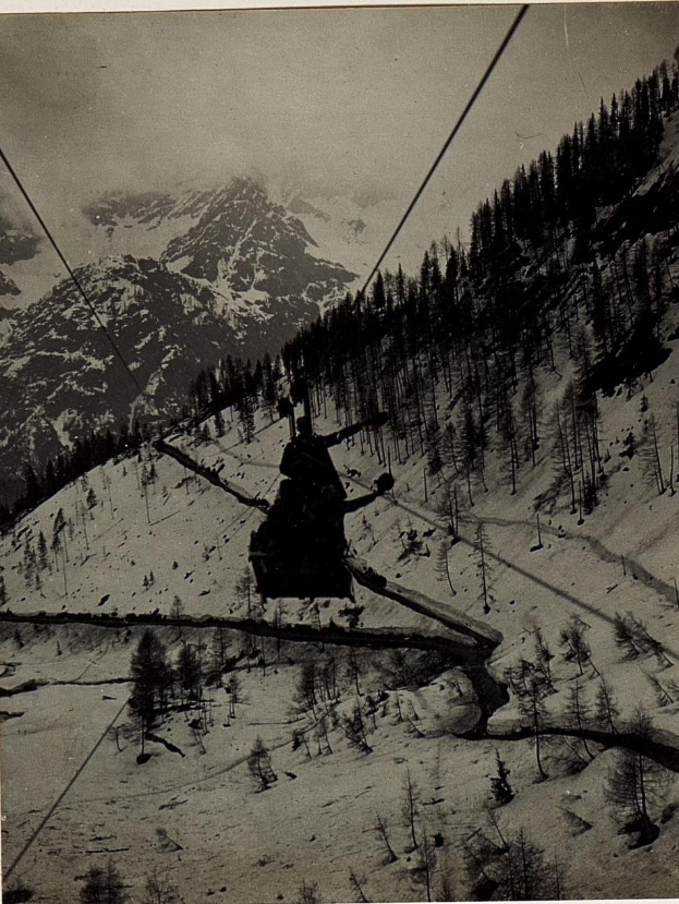 Schwarze und weiße Fotografie einer Person auf einer Skihebe mit schneebedeckten Bergen und Bäumen im Hintergrund und Text unten.