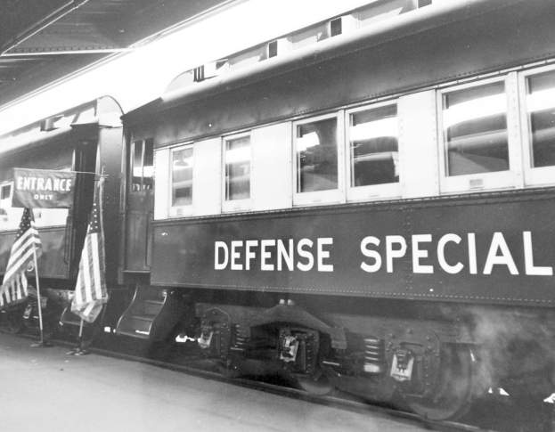 Schwarz-weiß-Foto eines Zuges mit der Aufschrift "Defense Special" auf den Eisenbahnschienen mit zwei Flaggen links und einer Decke oben.