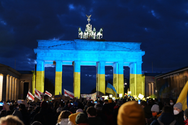 Eine Menge steht vor dem Brandenburger Tor in Berlin, hält Fahnen und Schilder, mit einer Protesttext tragenden Plane rechts.