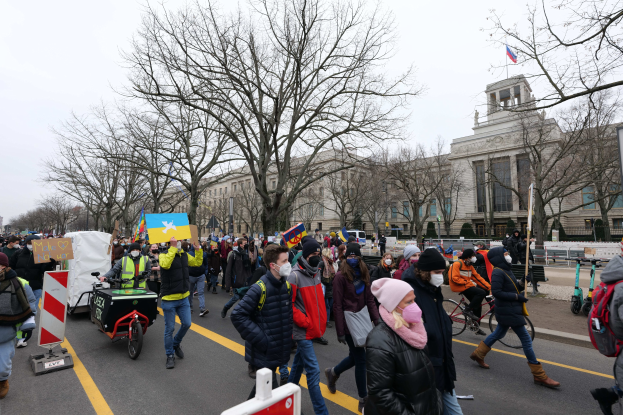 Eine große Gruppe von Menschen marschiert auf einer Stra\u00dke in Washington, D.C., mit Schildern, Bannern und Fahrrädern, mit Bäumen, Schildern und einem klaren blauen Himmel im Hintergrund.