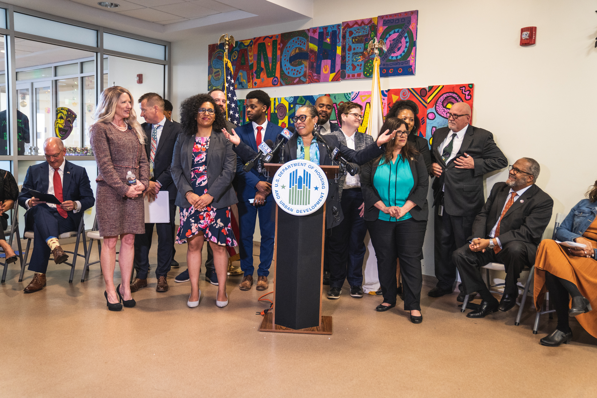 Eine Gruppe von Menschen, die vor einem Podium mit Mikrofon und einer Tafel mit Text darauf stehen und sitzen, mit Flaggen, einer Tafel, einem Feuerlöscher und Deckenlampen an der Rückwand.