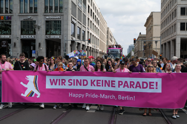 Eine Gruppe von Menschen geht auf einer Straße in Berlin, Deutschland, mit einer pinken Fahne, auf der 'Happy Pride March' steht, vorbei, mit Gebäuden, Laternenmasten und Verkehrszeichen an der Straße.