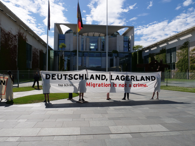 Gruppe von Menschen mit einem Transparent, auf dem "Deutschland, Lagerland Migration ist kein Verbrechen" steht, vor einem Gebäude mit Fenstern, umgeben von Gras, einem Metallzaun, Pflanzen, Bäumen und Fahnenstangen unter einem bewölkten Himmel.