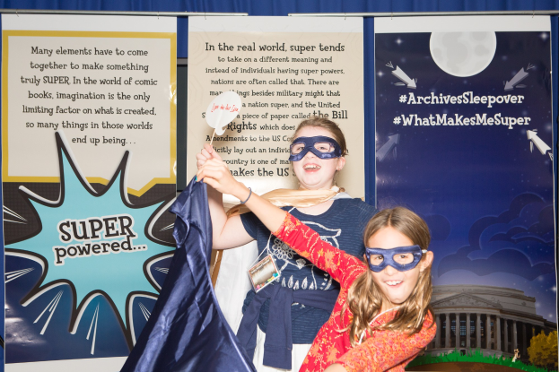 Zwei junge Mädchen in Superheldenkostümen posen für ein Foto vor einem blauen Vorhang bei einem Schulfest, eine hält ein Tuch, während beide Masken tragen.